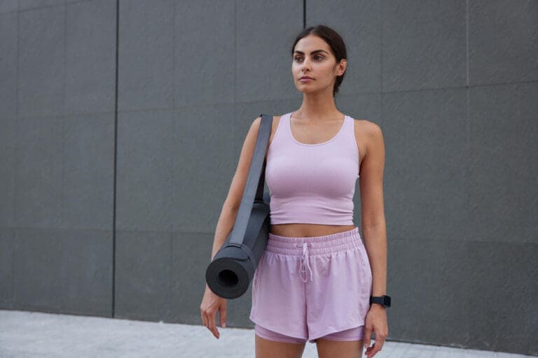horizontal shot motivated slim woman dressed sportswear prepares hard training poses with fitness mat focused into distance poses against grey wall with mock up space your text