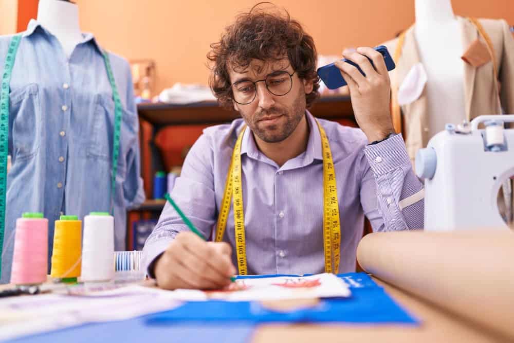 Do Sewing Patterns Include Seam Allowance? 2 young hispanic man tailor listening voice message by smartphone drawing clothing design atelier