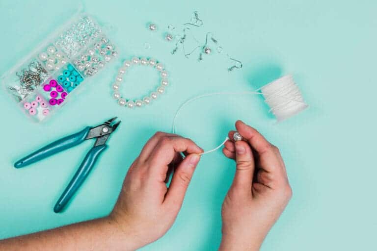 close up woman s hand inserting pearl white thread making bracelet teal backdrop